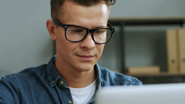 Close up portrait of handsome young business man in the glasses working on the laptop in the stylish office.