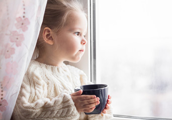 A little girl in the white knitted sweater sits on a window and drinks from a cup - 167429254