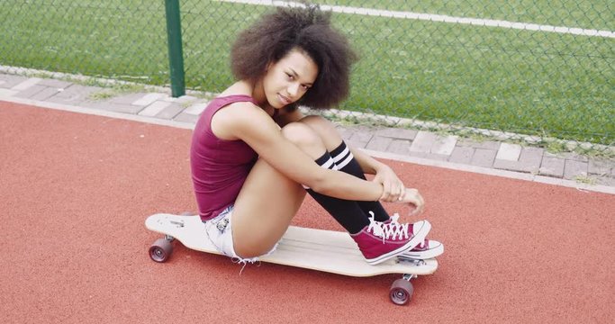 Beautiful young model in stylish clothing sitting on wooden longboard embracing knees and looking at camera smilingly.