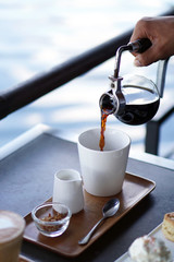 Barista pouring hot coffee from syphon coffee machine to white ceramic cup on wooden tray and blurred background, Selective focus.