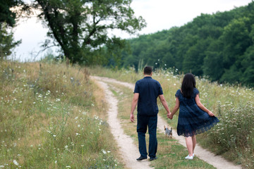 Fototapeta premium Couple in love holding hands and walking on footpath with yorkshire terrier dog outdoors near the forest in summer, back view. Man and woman walking yorkie dog outdoors, free space