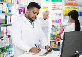 Chemist Writing On Clipboard While Colleagues Working In Pharmac