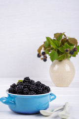 Composition of branch of Blackberry with leaf and  blackberries in a blue ceramic bowl on white table. 