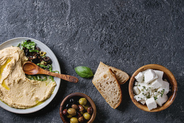 Homemade traditional spread hummus with chopping olives and herbs on blue plate, served with bread, olives, feta cheese, olive oil, spoon on black texture background. Mediterranean snack. Flat lay