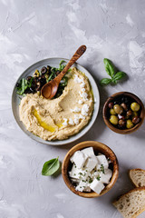 Homemade traditional spread hummus with chopping olives, oil and herbs on blue plate, served with bread, olives, feta cheese, spoon, on gray texture background. Mediterranean snack. Flat lay
