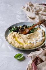 Homemade traditional spread hummus with chopping olives, oil and herbs on blue plate, served with feta cheese, spoon, basil, textile on gray texture background. Mediterranean snack. Close up