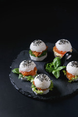 Mini rice sushi burgers with smoked salmon, green salad and sauces, black sesame served on slate stone board over black background. Modern healthy food
