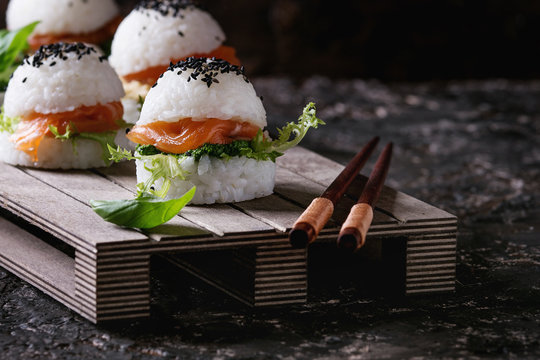 Mini rice sushi burgers with smoked salmon, green salad and sauces, black sesame served on wood pallet tray with chopsticks over dark brown concrete background. Modern healthy food. Close up