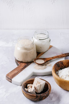 Rye and wheat sourdough in glass jars, fresh and instant yeast, olive wood bowl of flour for baking homemade bread. With spoon, serving board over gray concrete background.