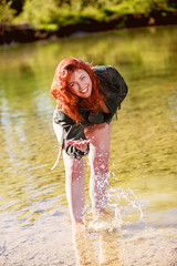 Rothaarige Frau mit Lederjacke steht im Wasser und spielt damit, mit Blick in die Kamera