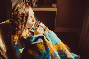 Winter cold concept. Young freezing woman in comfortable chair warming frozen hands wrapped in warm...