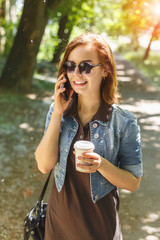 Young beautiful happy girl drinks coffee in the park on the phone on a sunny day