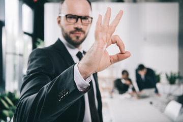 Fototapeta na wymiar handsome young businessman showing okay sign with blurred colleagues on background
