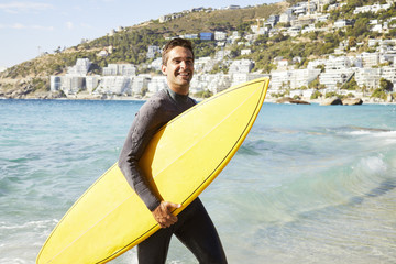 Happy surfer in wetsuit carrying surfboard on beach