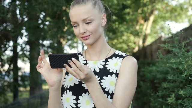Pretty girl in floral shirt smiling to the camera while using smartphone in the park, steadycam shot
