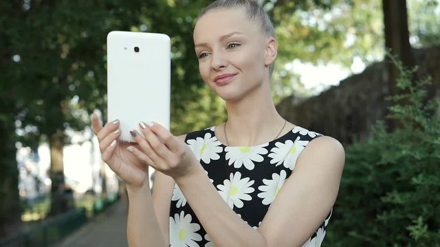 Blonde girl in floral shirt standing in the park and browsing internet on tablet, steadycam shot

