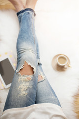 Fototapeta na wymiar A young woman wearing distressed jeans sitting on wood floor on a white fur carpet at home. Around a cup of coffee, tablet and notebooks. Gold bright feminine theme