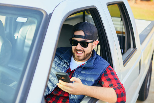 Handsome happy men driver with a beard smiling in the pickup car truck, looking at mobile phone, searching information in Internet, using social media. Attractive male driving big vehicle in hat.