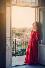 Portrait of beautiful young woman with Curly long hair style in long red dress staying near balcony, enjoy sunny day in Europe travel