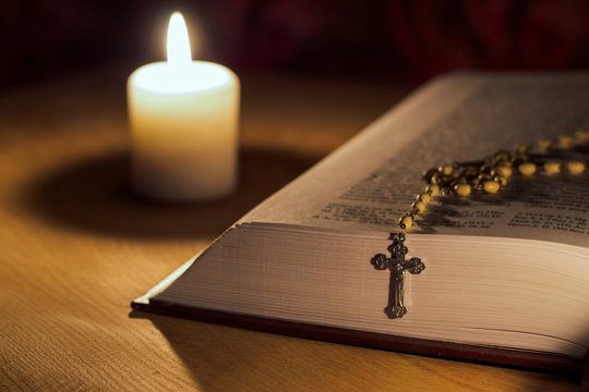 A burning candle, a bible and a cross a rosary on a wooden table