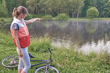 Young female biker next to her bike looking at the lake in a park and showing to something on it.