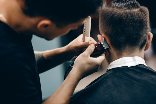 Close-up of a young hipster guy in a barber shop hairdresser cutting hair with scissors and a typewriter, waxing hair, blow-drying. Concept men's place, toned photo. Soft focus.
