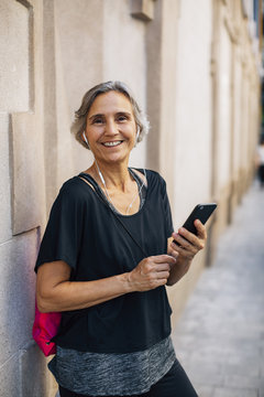 Portrait Of Happy Woman Holding Smart Phone While Standing Against Wall