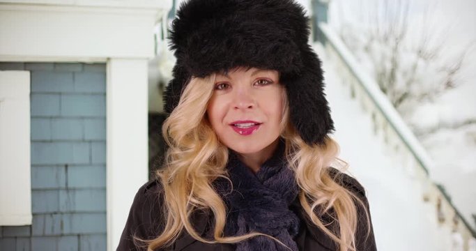 Cheerful Caucasian woman in cozy hat and scarf standing outside home in the snow smiling at camera. Close up of attractive white female in furry cap enjoying the winter season outdoors. 4k 