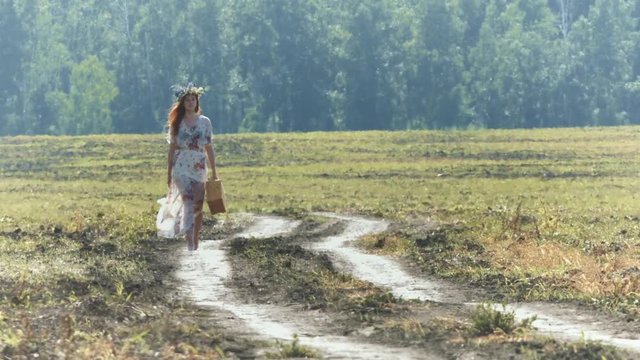 Walk in the field.

A beautiful red-haired girl with a wicker suitcase walks along the country road in the field. Slow motion.