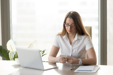 Attractive businesswoman holding smartphone, texting message while sitting at workplace, using apps, banking application, person dialing number, adding reminder, important note on phone, mobile office