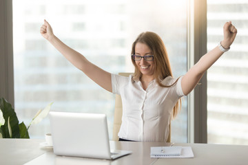 Excited smiling businesswoman celebrating business success at workplace, raising hands looking at...
