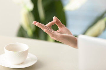 Female hand in chin mudra yogic gesture, peaceful calm woman practices meditation at home office desk with laptop and porcelain cup, yoga at work for relaxation, exercises to reduce stress, close up