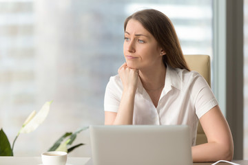 Discontented thoughtful woman with hand under chin bored at work, looking away sitting near laptop,...
