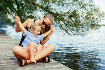 Group portrait of happy white Caucasian mother and daughter child having fun outside. Mom and child girl playing, hugging in park on pier by water lake. Candid authentic real lifestyle. - obrazy, fototapety, plakaty