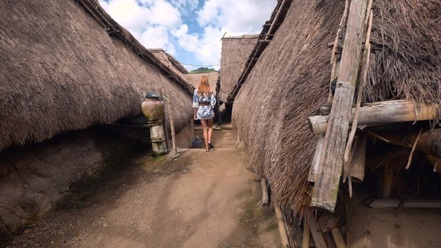 Young caucasian woman walking on street in Sasak Sade traditional weaving village in Lombok with bamboo houses and grass roofs. Shot with Sony a7s and Atomos Ninja Flame on sunny day with blue sky.