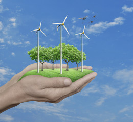 Fototapeta na wymiar Wind turbines, grass and trees in human hands over blue sky with white clouds, Ecological concept