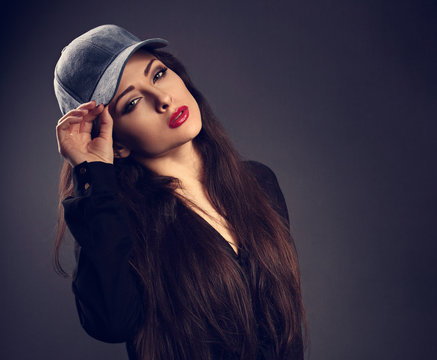 Beautiful sexy young make-up model in blue baseball cap with red lipstick posing on dark shadow grey background holding the cap. Toned closeup portrait