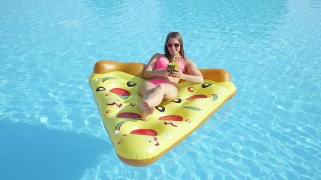 SLOW MOTION CLOSE UP: Happy brunette girl sipping pineapple drink and relaxing on inflatable pizza float. Young woman in bikini enjoying summer vacation drinking cocktails on fun pizza floatie in pool