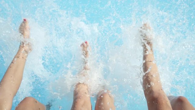 SLOW MOTION CLOSE UP: Three unrecognizable cheerful girls sitting on pool edge, splashing water with their feet on hot sunny day. Three playful young women on summer vacation splashing water with legs
