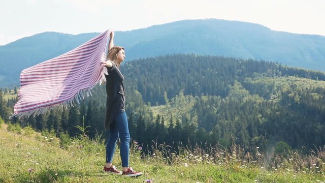 Young woman standing high top mountain looking forest holding blanket above head wind blowing flying windy summer happy vacation wild freedom carefree satisfied back full view breathing fresh air peak