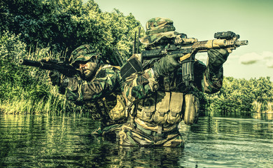Pair of soldiers in action during river raid in the jungle waist deep in the water and mud and...