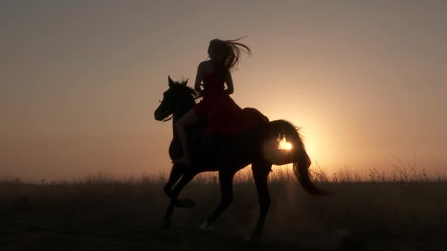 Young girl in red dress riding black horse against the sun. Silhouette of woman rider with her stallion galloping across a field kicking up dust at sunset. Horseback riding in slow motion.