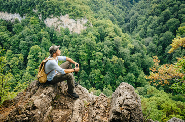 Naklejka na ściany i meble Hiker, sitting on a rock climbing to the height of a bird's flight and examining the tops of trees. View of the forest from the top. Travel carrying things in a backpack.