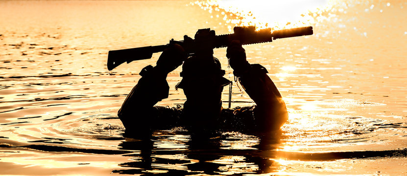 Silhouette of special forces with rifle in action during raid crossing river in the jungle waist deep in the water, with weapons over his head