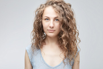 Close up shot of tender and cute teenage female with big blue eyes, long curly hair and clean heathy skin looking straight at the camera while posing indoors against white studio wall.
