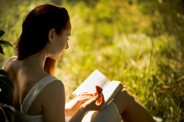 Rear view of attractive woman sitting on grass with book in her hands. Young foxy haired woman...