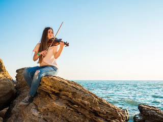 Young beautiful caucasian girl wearing jeans plays violin staying on the beach near the beautiful...