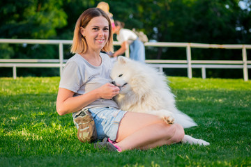 An adult woman with red hair plays and strokes her dog of the Samoyed breed. White fluffy pet in a park with mistress on a green lawn have fun.