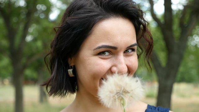A young woman takes a dandelion before her mouth and smiles sincerely in slow motion. She frolicks and looks around being on a picturesque lawn with green trees in summer