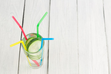 Many slices of green fresh cucumber in glass with water and four colorful plastic tubule on old white wooden planks